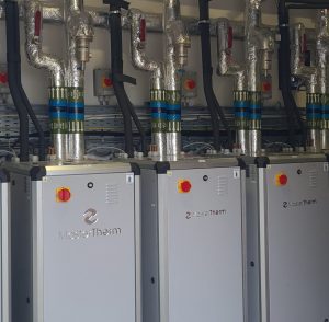 Row of retrofitted industrial heat pump units with insulated pipework and control panels in pump room.