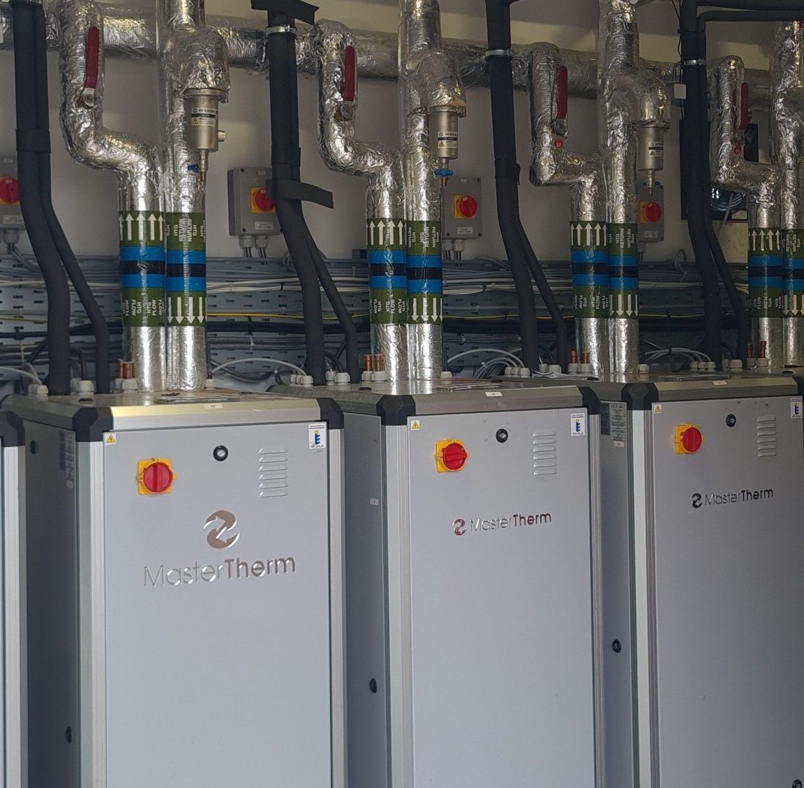 Row of retrofitted industrial heat pump units with insulated pipework and control panels in pump room.